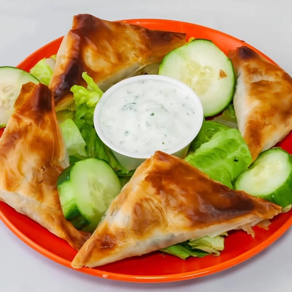 Four golden-brown phyllo pastries, likely spanakopita, with a side of tzatziki, lettuce, and cucumber slices.