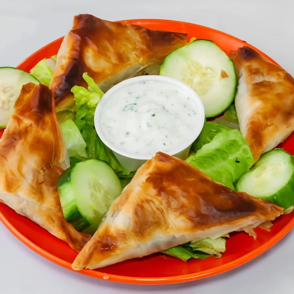 Four golden-brown phyllo pastries, likely spanakopita, with a side of tzatziki, lettuce, and cucumber slices.