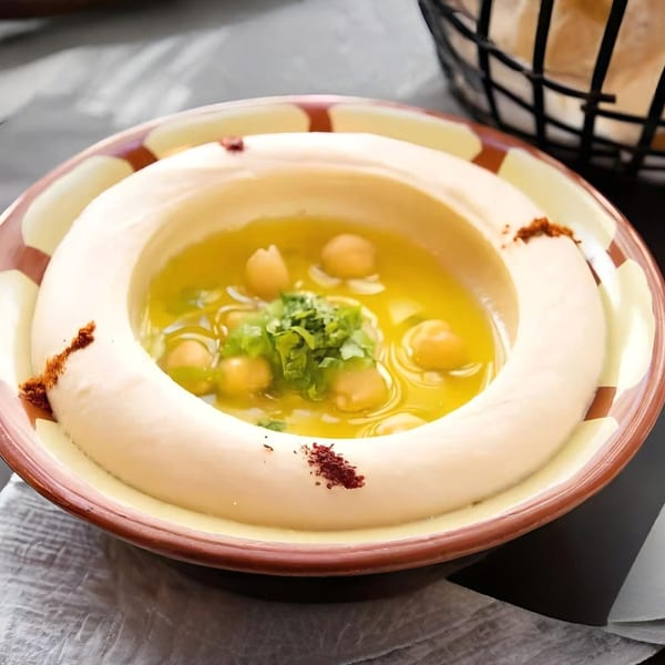 A bowl of KINZI hummus with olive oil, chickpeas, and parsley, next to a bread basket.