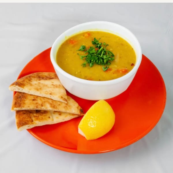 A bowl of Kinzi's Mediterranean lentil soup with pita bread and a lemon wedge on an orange plate.